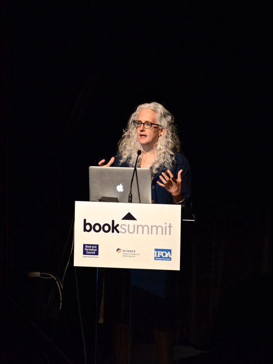 Image of Kate giving a talk at an event, standing in front of lectern with a mic and a laptop in front of her, with a black background. Kate is wearing a dark blue shirt.