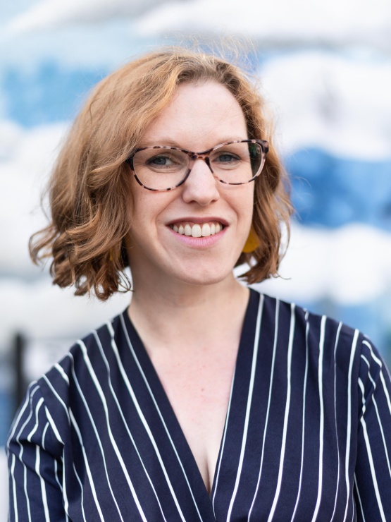 Image of Verity smiling into the camera, wearing a dark blue and white striped top, with red hair and tortoiseshell glasses