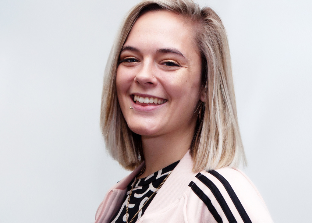Image of Grace looking at the camera and smiling. She has blond hair and is wearing a pink adidas top and printed black tee.