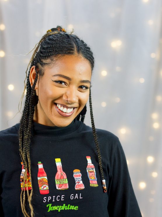 Josephine Gyasi smiling at the camera. She has long black braids and is wearing a black hoodie with cartoon drawings of hot sauce on it and the words 'Spice Girl' underneath.