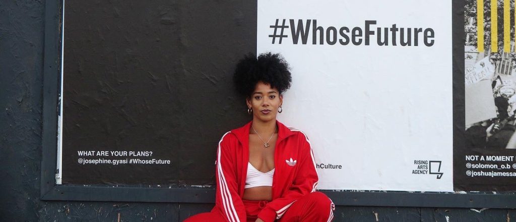 Image of Josephine Gyasi outside, looking at the camera with attitude, beside a billboard that asks 'What Are Your Plans? She is wearing a red adidas tracksuit and white crop top.