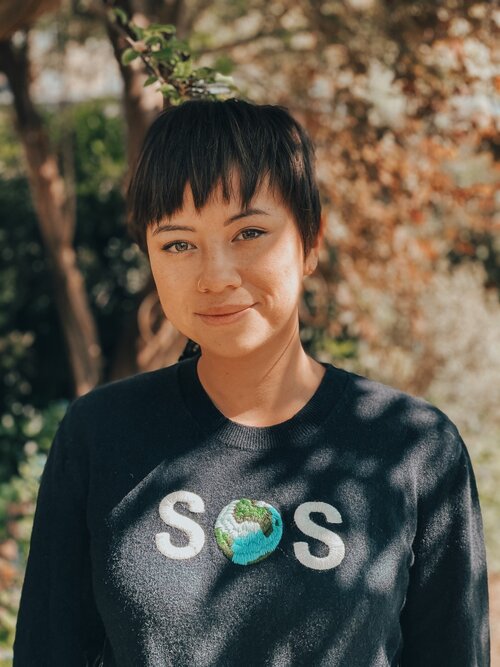 Image of Tori Tsui smiling at camera, outside in natural light, with a jumper that says 'SOS'. The 'O' in SOS is a picture of the planet.