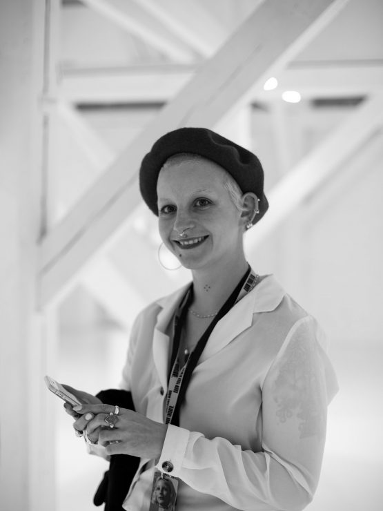 A picture of Tessa, a young white woman photographed in black and white wearing a beret and smiling at camera. Tessa has multiple face piercings.