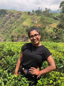 Young woman with black hair tied up, black t-shirt, standing in front of lush green field and hills