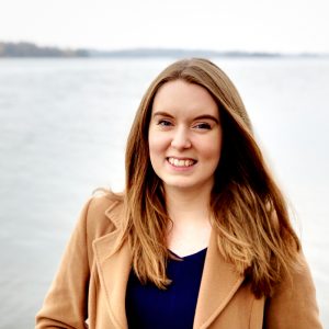 White woman with mid/long light brown hair and a camel coat smiling at camera