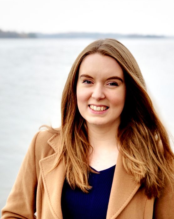 A picture of Jenny, a white woman with mid/long light brown hair and a camel coat smiling at camera