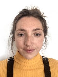 A headshot of Jess, a white woman with brown hair tied up and a nose piercing. Jess is wearing a yellow roll-neck top and black dungarees.