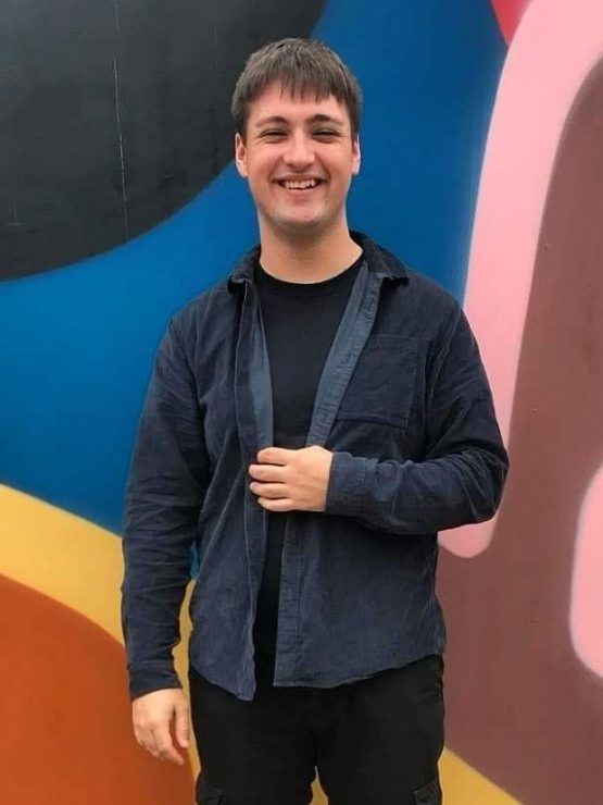 An image of Jack who is smiling into the camera in front of a colourful background. Jack is a white man with short brown hair. He is wearing a black top, blue cord overshirt and black cargo trousers