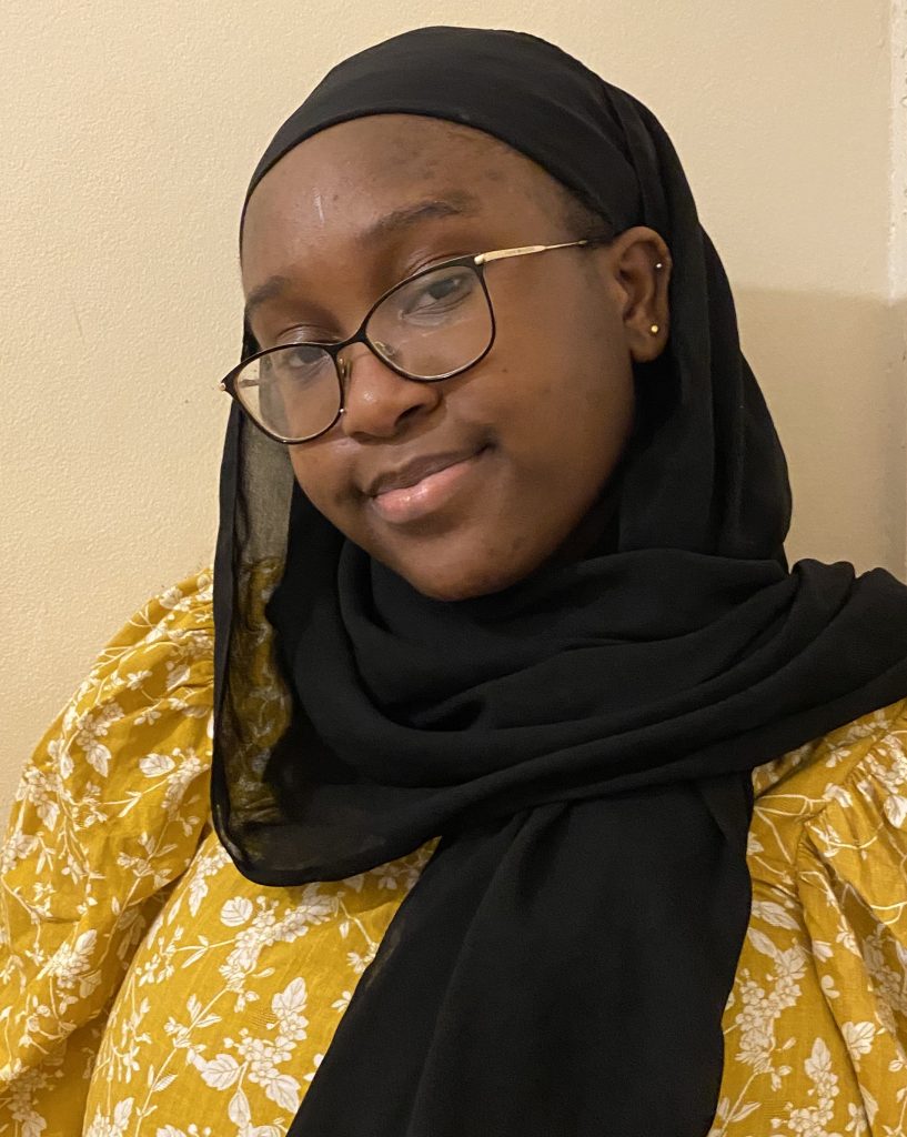 A headshot of Bashirat who is smiling into the camera in front of a pale background. Bashirat is wearing a yellow floral top, black headscarf and has black and gold glasses.