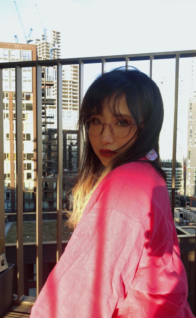 A image of Vivienne who is staring into the camera with sky scrapers behind her. She has long brown hair and pair of round glasses with pink frames. She is wearing a pink jumper.