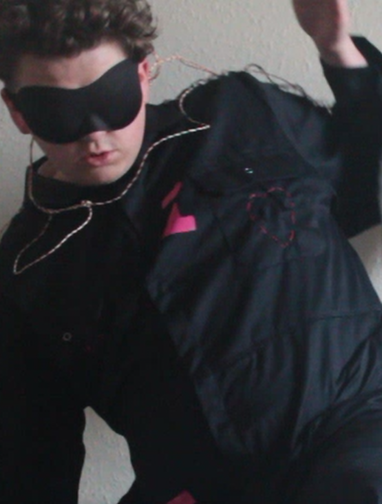 A headshot of Oma who is looking away from the camera in front of a pale background. Oma is wearing a black boiler suit with pink embroidery. Oma has an eye mask on and ear plugs and is moving quickly in the image.