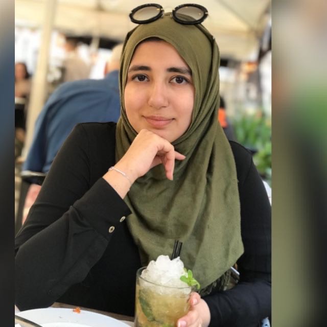 A woman wearing a green hijab is smiling as she drinks a cold drink with lots of ice