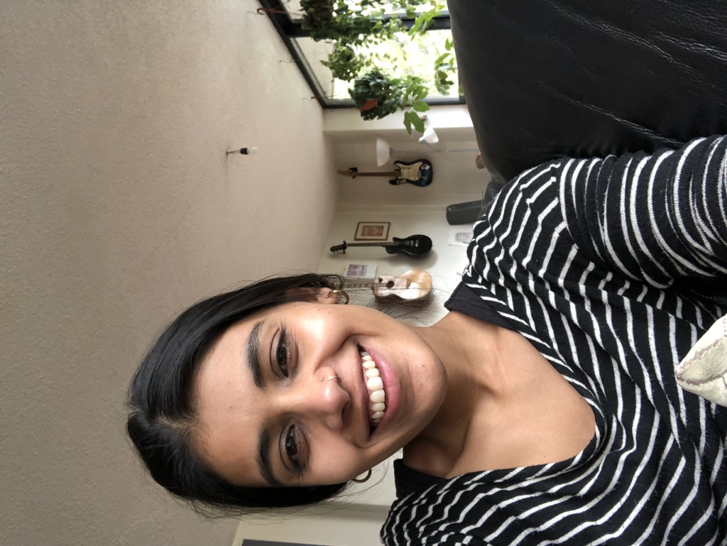 A woman with dark hair tied back and a black and white striped top is taking a selfie indoors and smiling