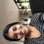 A woman with dark hair tied back and a black and white striped top is taking a selfie indoors and smiling