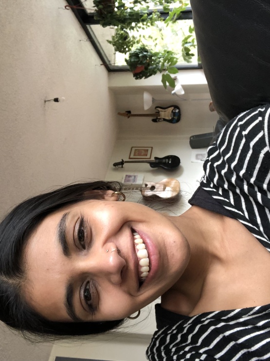 A woman with dark hair tied back and a black and white striped top is taking a selfie indoors and smiling