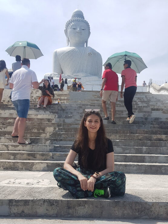 A young woman is sitting on stone steps below a large stone Buddha