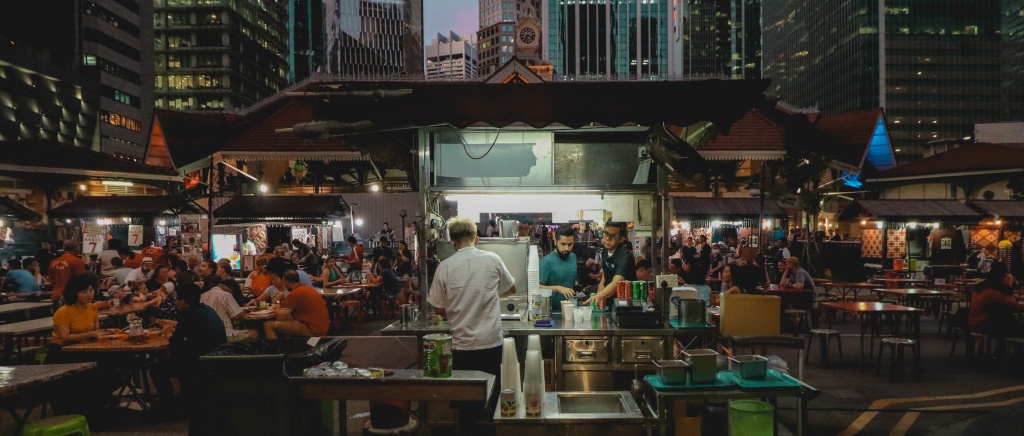 A hawker market in Singapore at dusk, with lights on, people sitting down at tables to eat, and people cooking street food dishes at small stalls