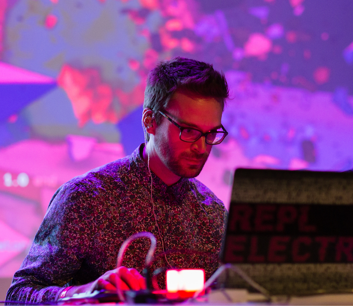 A white man with short brown hair, glasses and a short beard is using a laptop computer at an event lit with pink and purple lights