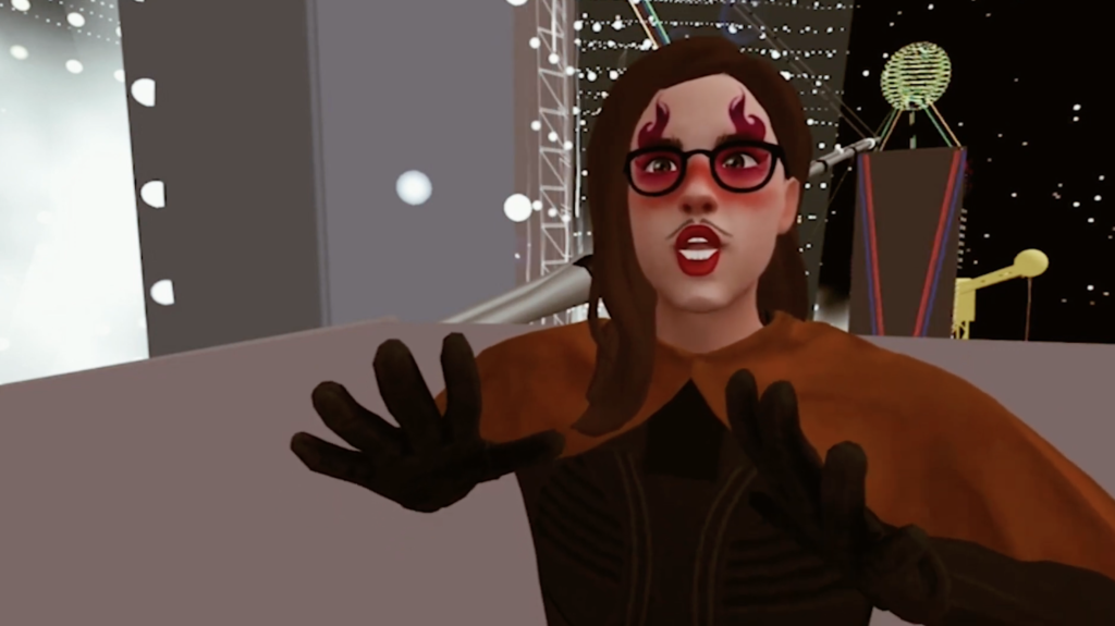 A virtual reality character holds out their hands. They wear an orange cape and have long dark hair. They are wearing glasses, and have red makeup almost like flames rising up the sides of their face