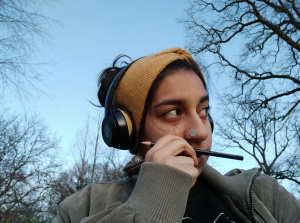 A photograph of an Indian woman with a large flat silver nose stud. She is outdoors on a winter day, with trees behind her on a pale blue sky. She wears a green coat, a yellow knitted headband and headphones.