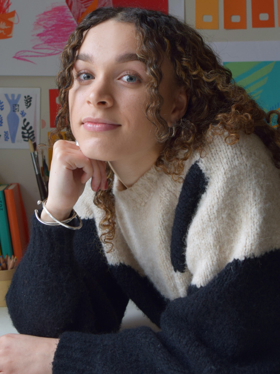 A phograph of a young woman with curly brown hair leaning on her desk, wearing an oversized black and white jumper
