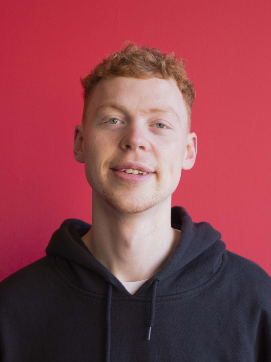 A photograph of a white man with ginger hair and blue eyes, wearing a black hoodie. He stands in front of a bright red wall.