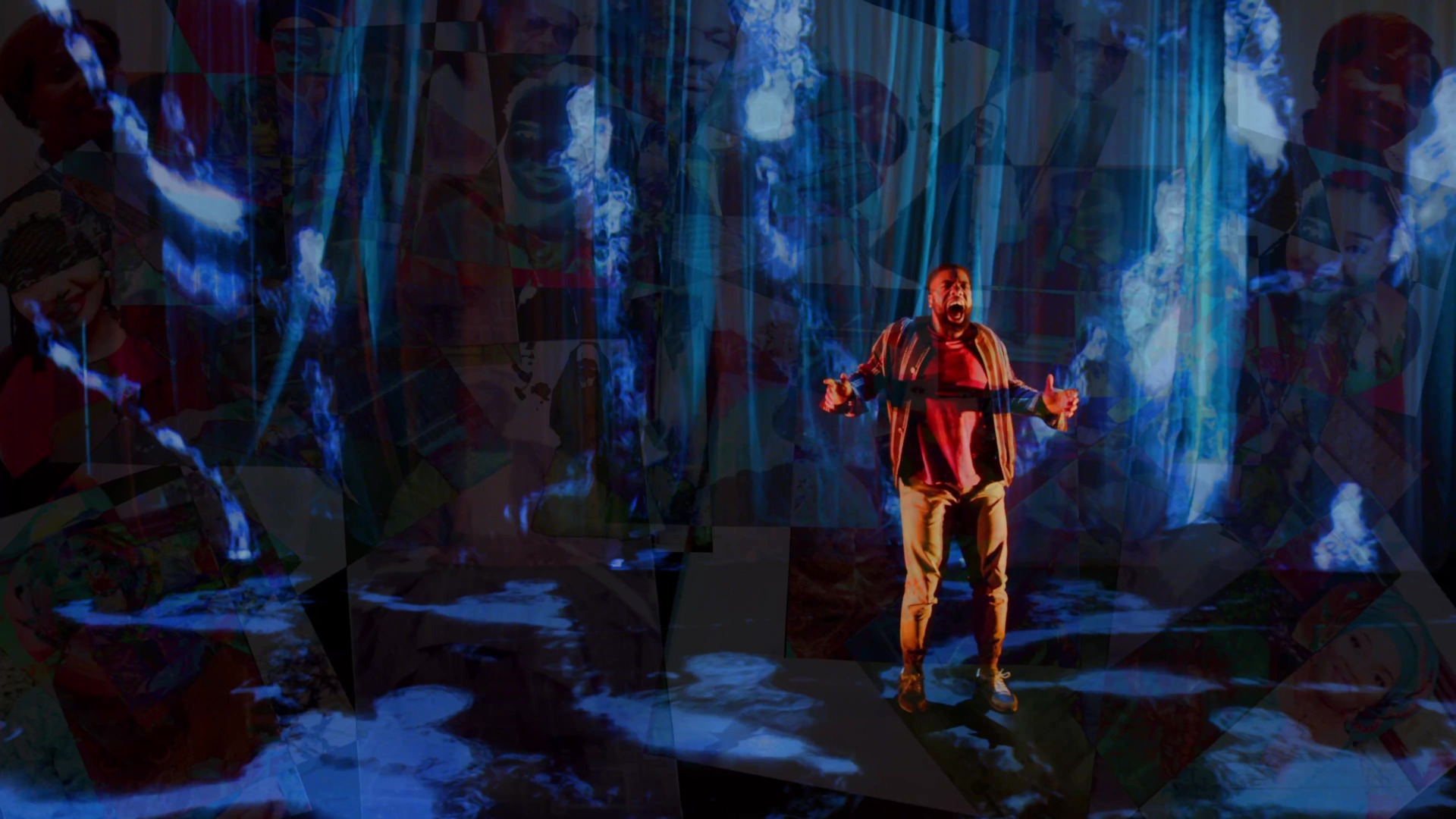 Image of young black man, hands out in front, yelling or screaming. He is standing on stage in front of a misty black backdrop with various projections. 