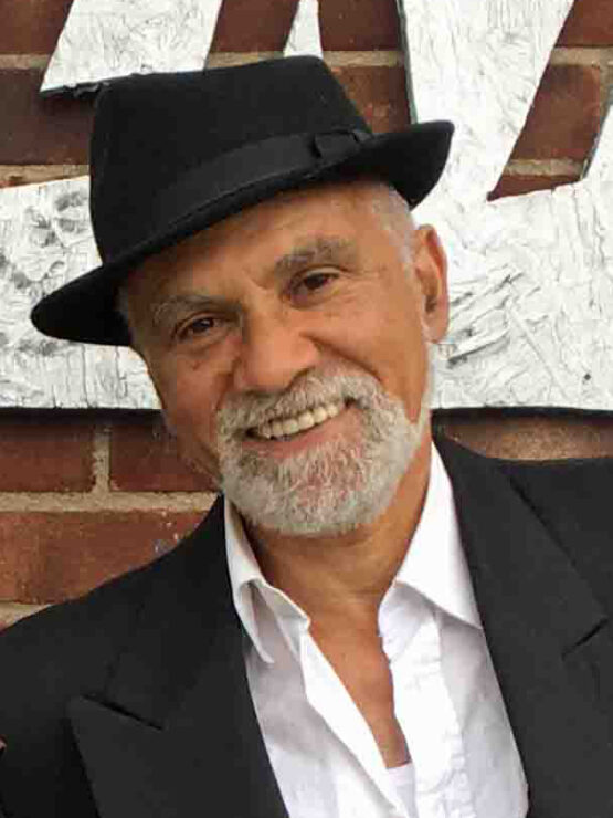 Older man with short white beard and black rimmed hat, open white shirt and black jacket in front of a brick wall