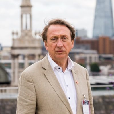 White man wearing cream jacket and white shirt standing in front of city scape