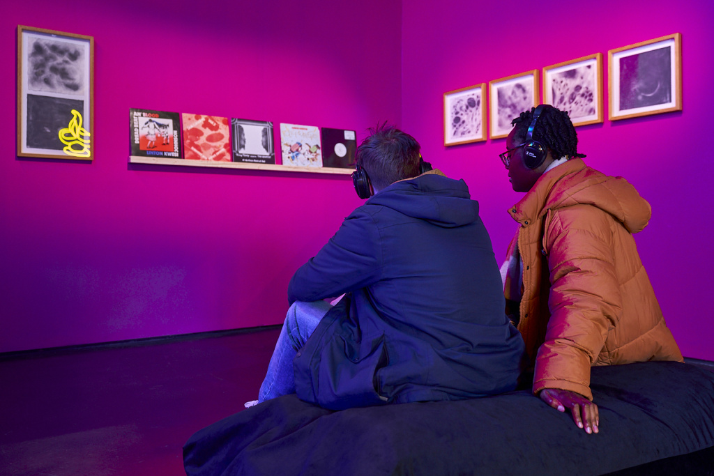 Two people sat on beanbag with earphones in, shelf of vinyl covers, and framed images in the background, in a purple room