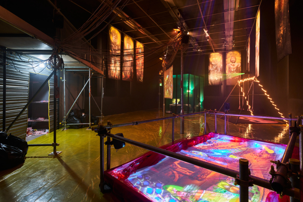 Exhibition view, a screen on the ground in a play pool with metal railing around it foregrounded, in the background a swing, glass living room and entrance to a dark covered area, with large vertical hanging banners featuring cgi images