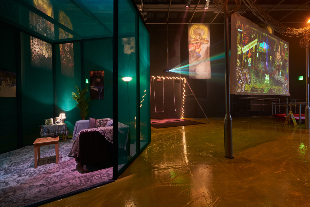 Exhibition space with a mock up modern living room in a glass walled space, with a swing set in the background, a lamppost in the foreground and banners with CGI figures in a church stained glass style, a projector shines a light off to the side