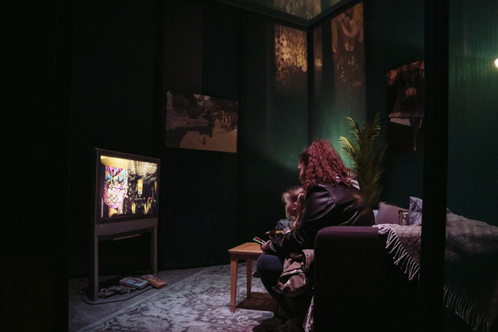 A woman and child sat on a couch in exhibition living room installation looking at television with CGI images.