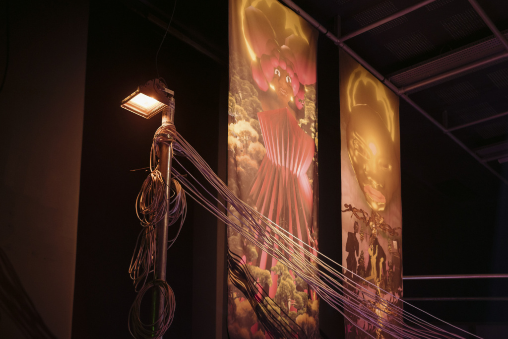 Exhibition, view of two large vertical banners on wall with cgi figures in a church stained glass/idol style and an active lamppost in the foreground with wires tied about the top
