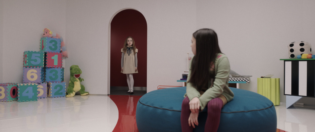 shot from M3GAN, a sad girl looks round to see a lifesize doll in a red archway in a white room full of toys