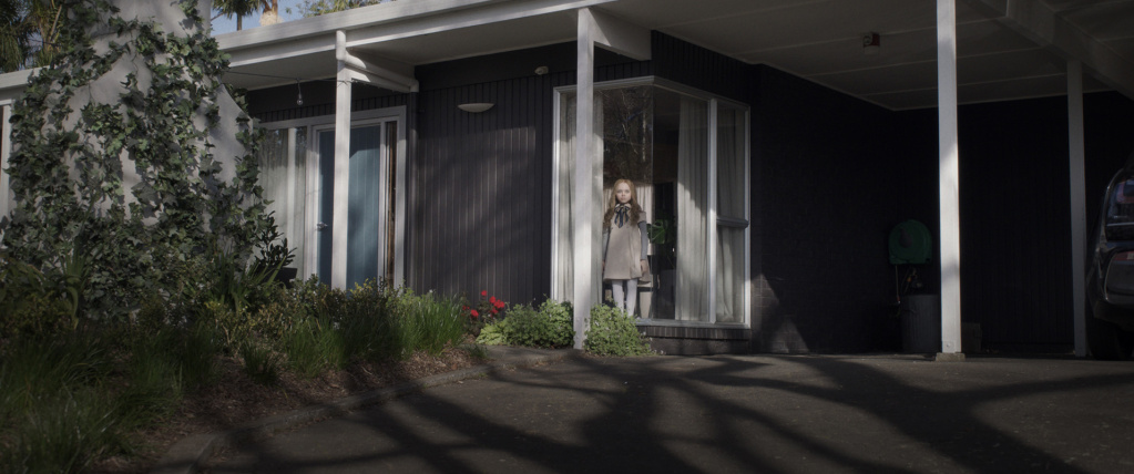 Shot from M3GAN, a lifesize doll stands on the porch of a modern suburban US home in a leafy area