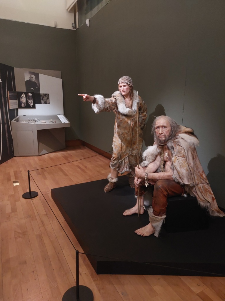 A display of two waxwork figures of early modern humans, one pointing, one sitting, both wearing animal hides. with a museum cordon around it