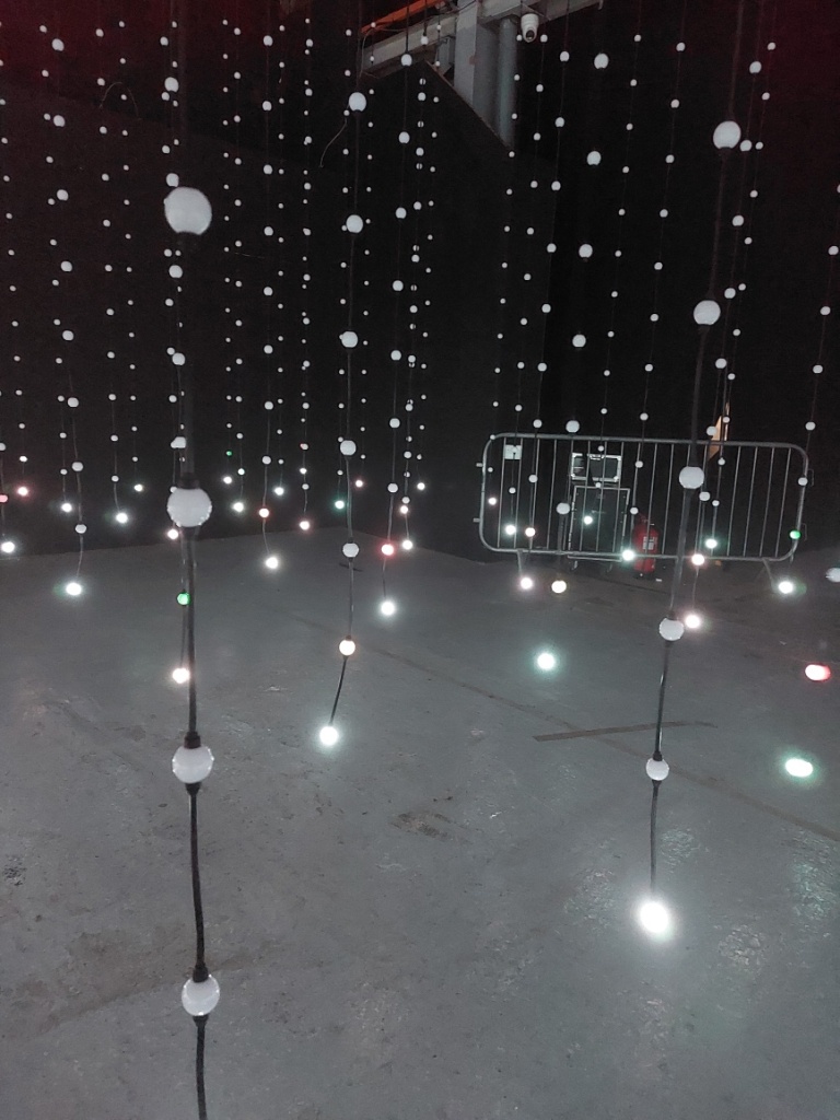 A view of dozens of lines of ceiling length light strings, glowing various colours at their base, in a dark room