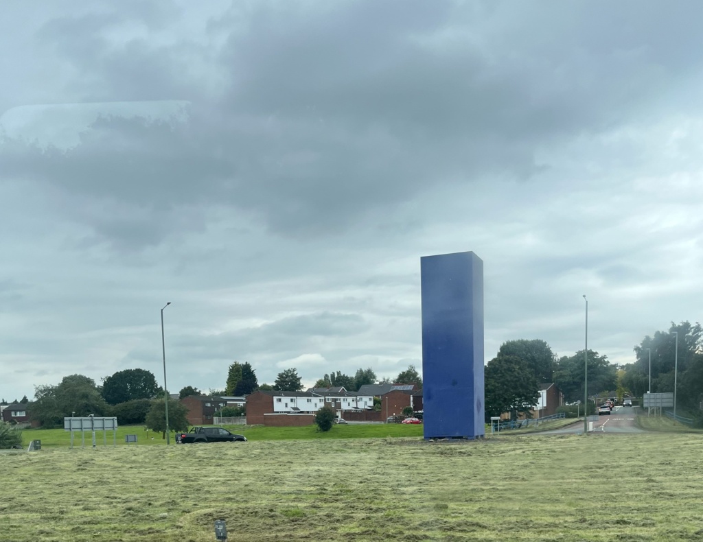 a blue box monolith on a grassy mound by a road on a cloudy day
