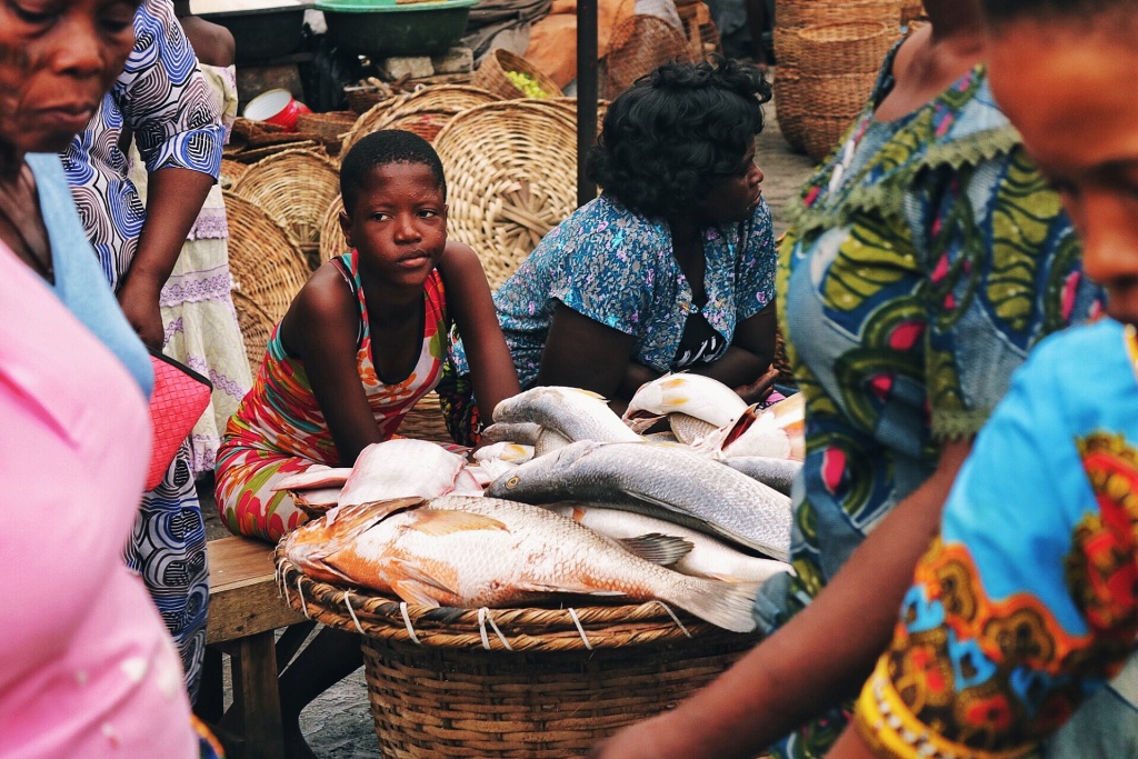 a fish stall at a market a girl and a woman sit behind an array of fresh fish resting on a wicker basket as people walk past