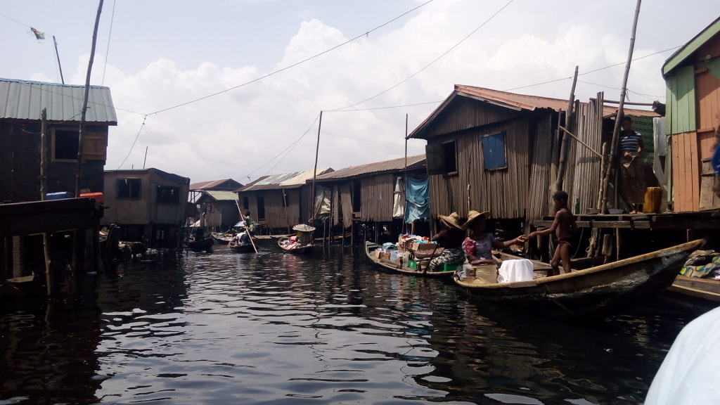 A canal street in Makoko, with shacks on poles raised above the water and vendors in boats on the edge of the water 
