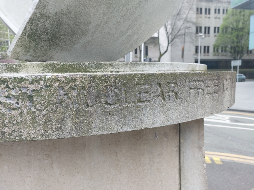 the plinth of a sculpture in a city area - the words 'nuclear free city' are visible but decayed