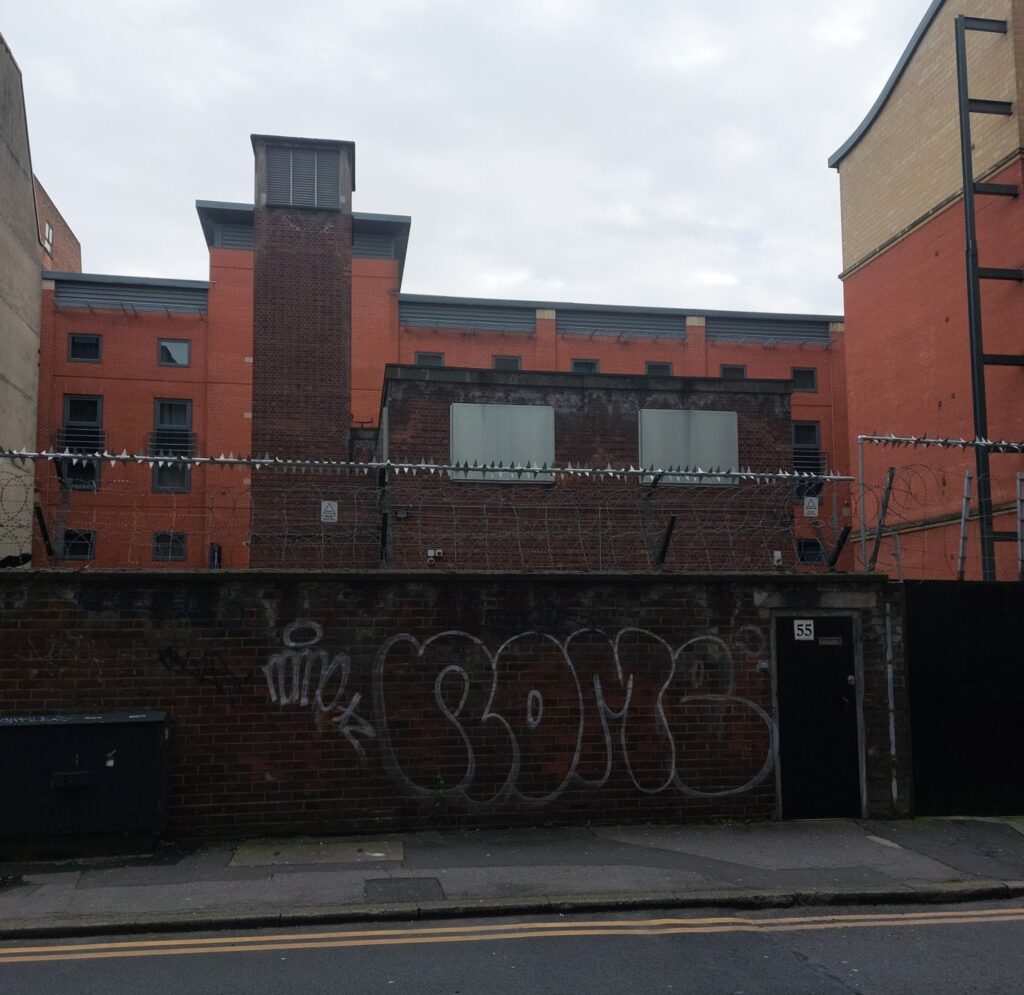 a redbrick building with a tower, obscured by a wall with barbed wire