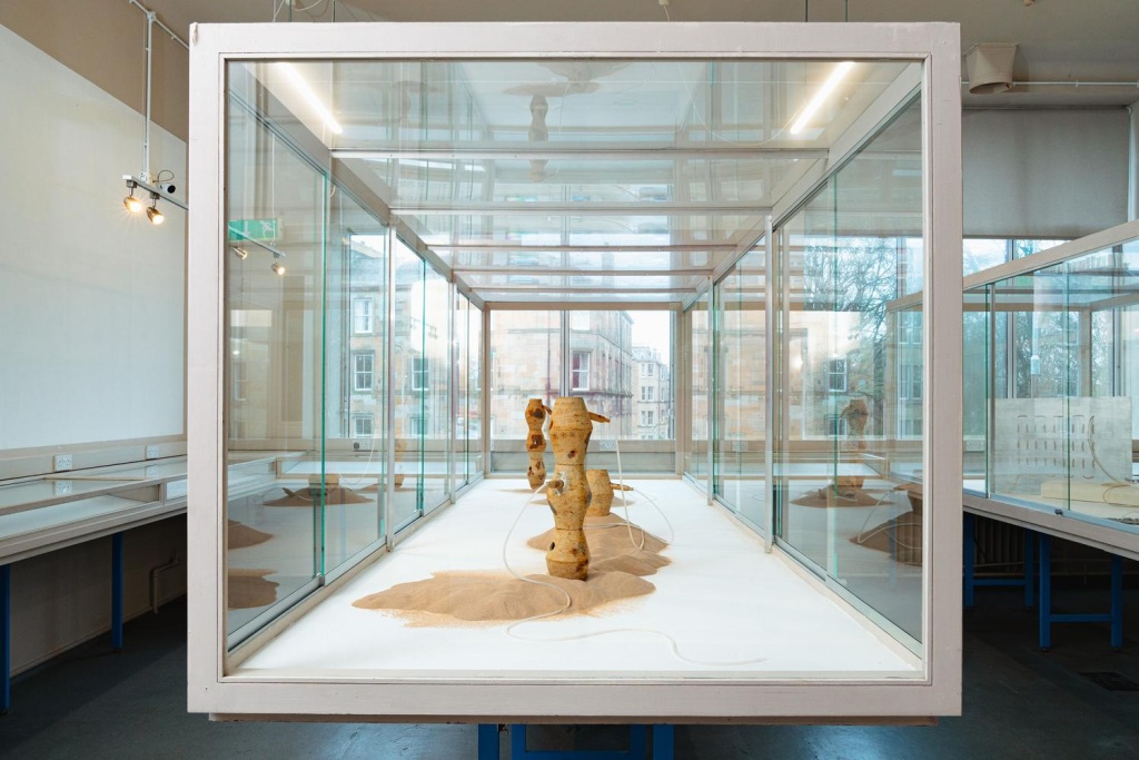 A sideview of a gallery display case featuring pots embedded in sand with tubes emerging from them