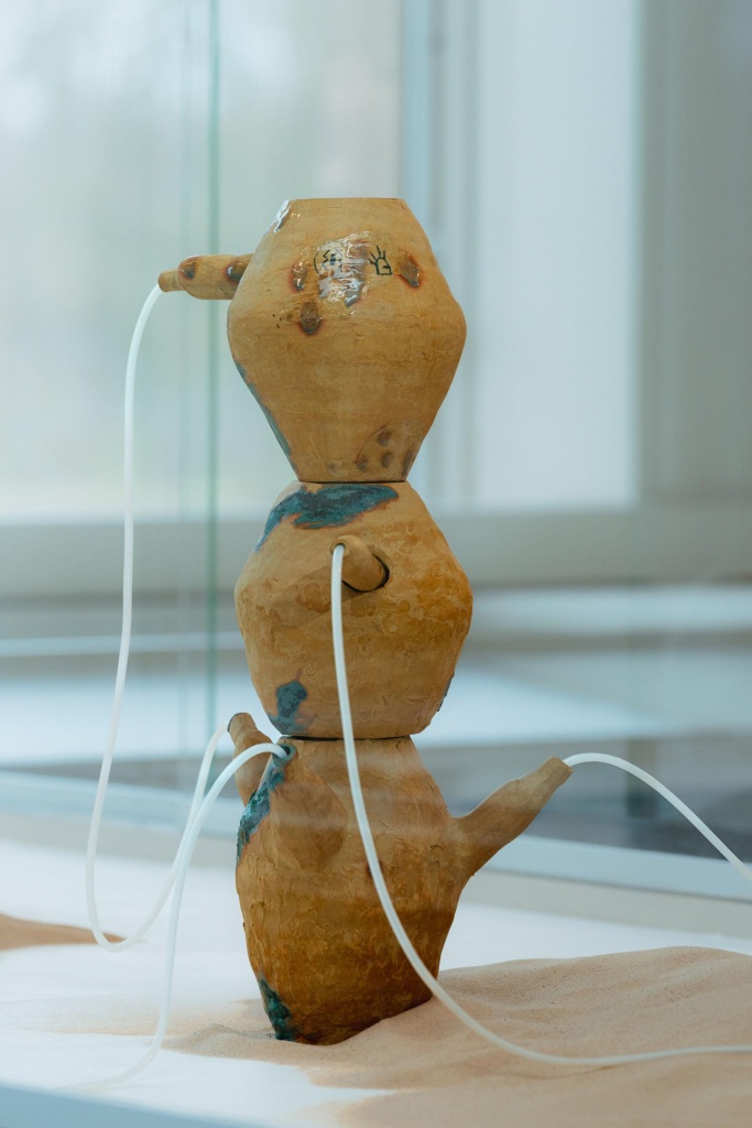 A sculpture with a ceramic tower with spouts that have tubes coming out, it stands in sand