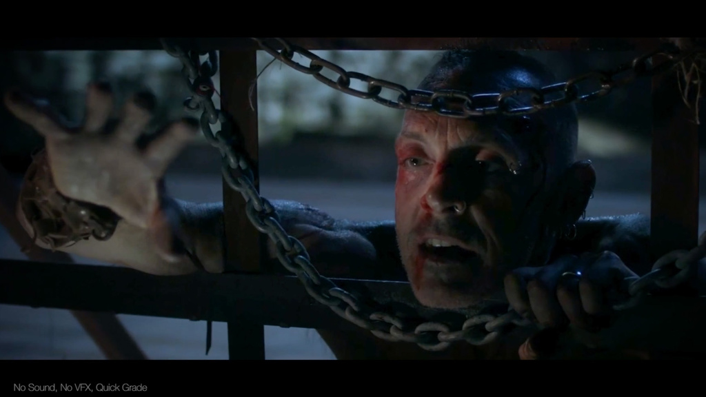 a movie scene, a person reaches through cage bars and chains in a desperate way, they have blood on their face