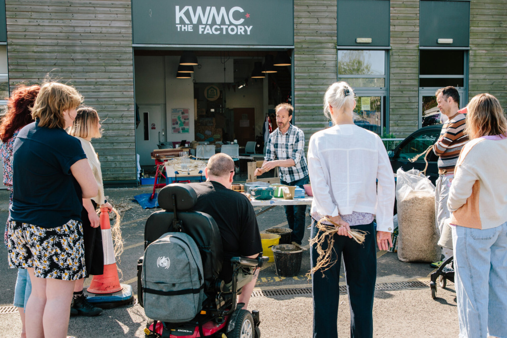 a group of 7-8 people arranged outside Knowle West Media Centre in an open courtyard watching someone at a table with a bunch of materials on presenting