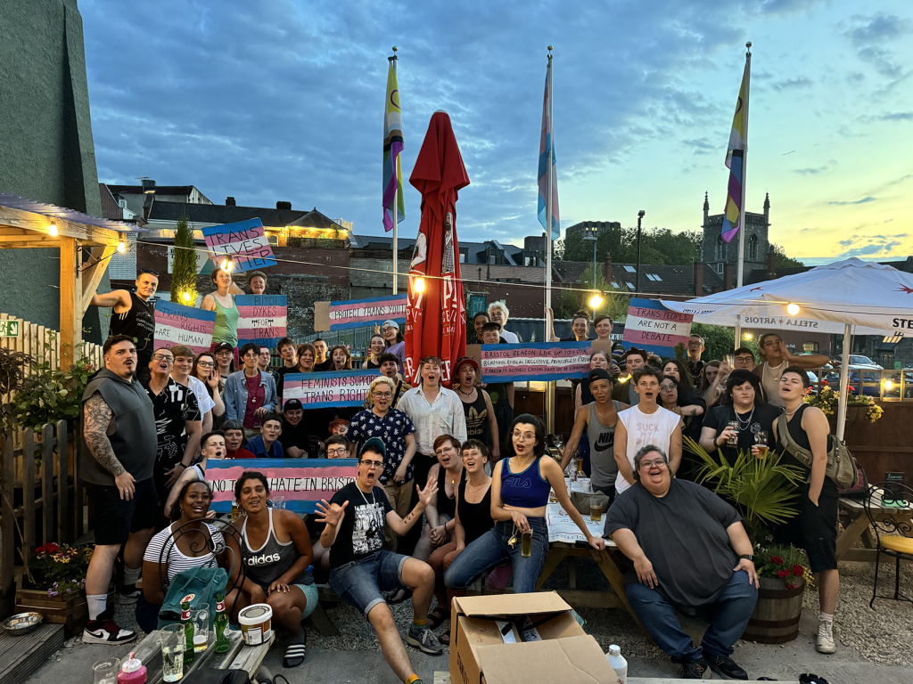 large group of butch bar visitors in a beer garden with signs supporting trans rights